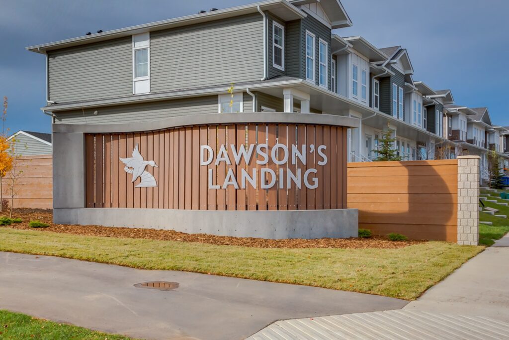 Dawson’s Landing neighbourhood entrance sign with a goose logo, wooden slats, and concrete framing, set in front of townhomes.