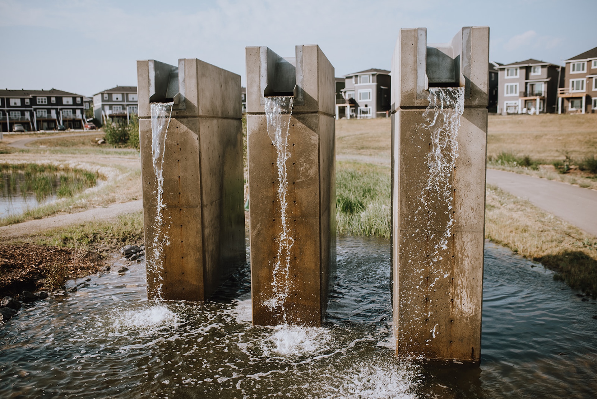 Three vertical concrete columns with water cascading from metal spouts at Dawson's Landing, with houses and a pond in the background.